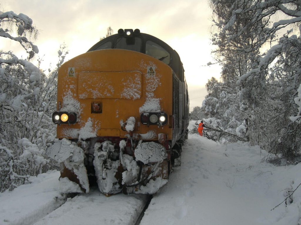 A small snow plough on the front of a locomotive, clearing snow off the tracks with a Network Rail worker in the background moving a tree
