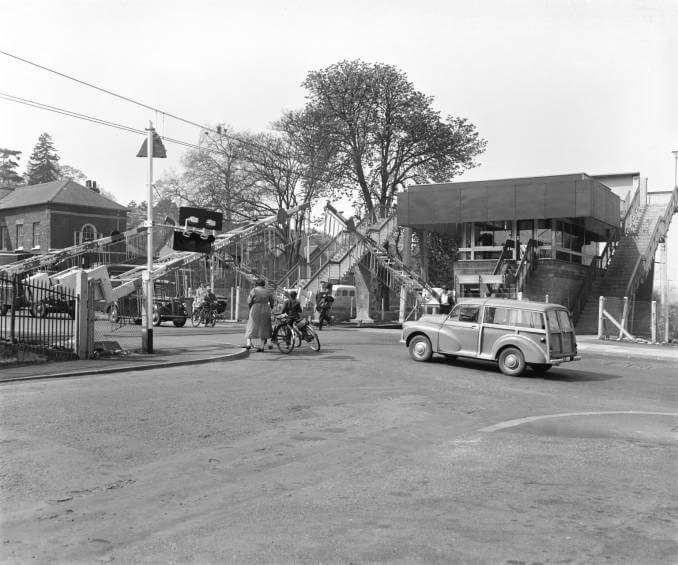 old photograph of a level crossing with barriers going up and a pedestrian, a bicyclist, and a motorist waiting to cross
