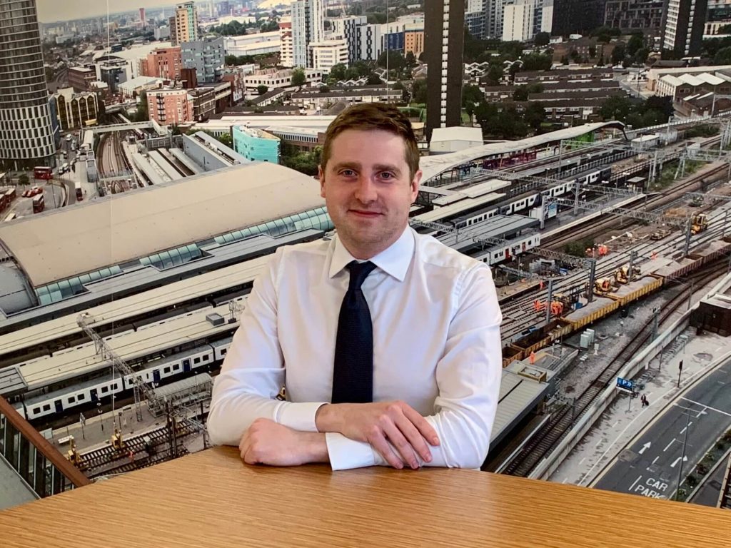 Owen Flanders, a former apprentice, sitting at a table in front of a large image of a railway station