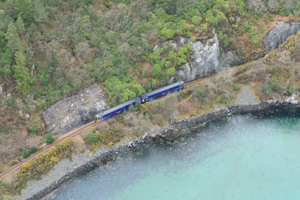 Aerial shot of a train traveling beside a rock face and water, daytime