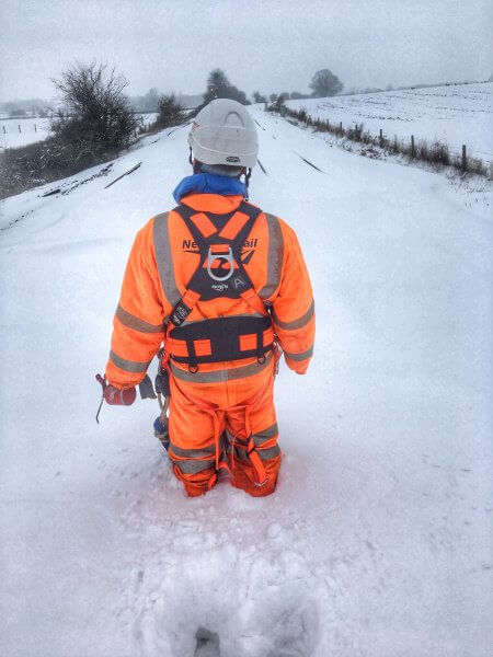 A railway worker in PPE stands in the middle of the tracks in a rural area with snow up to his knees, daytime