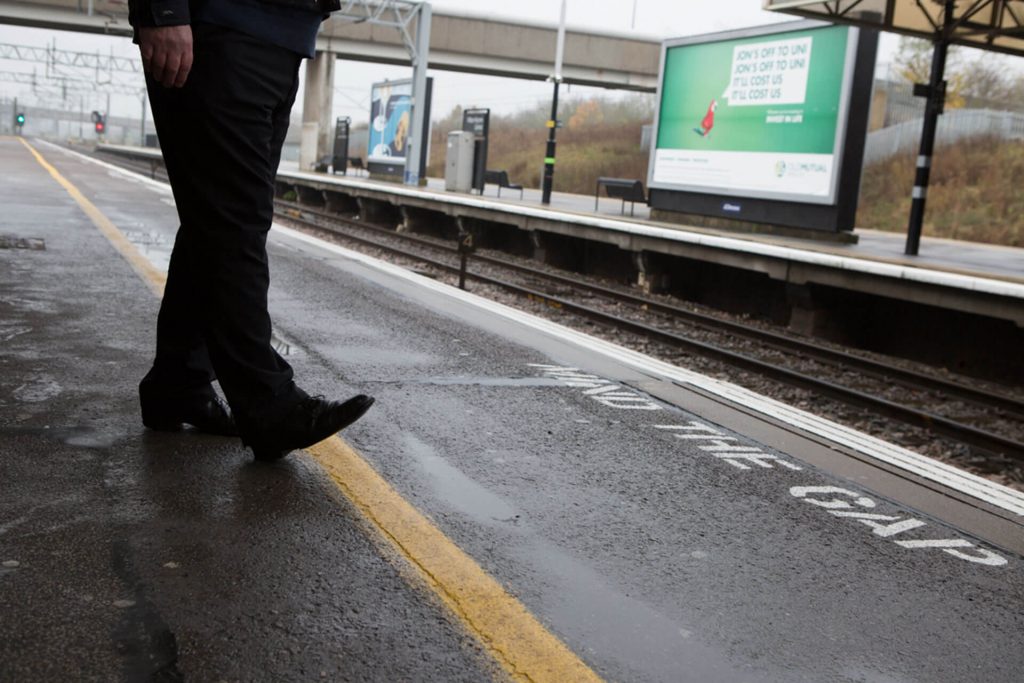 A close up photograph of someones feet on the yellow safety line at a train station platform.