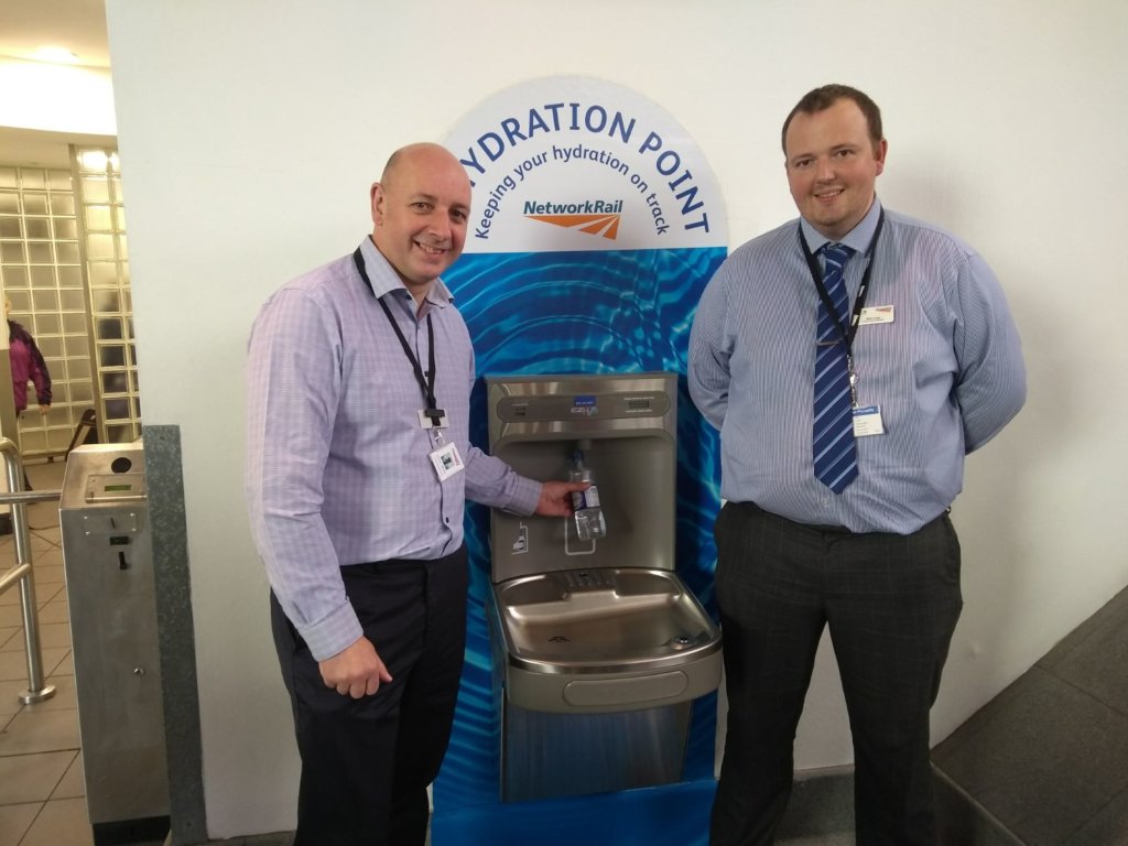 Two members of Network Rail staff using a free water fountain at a station