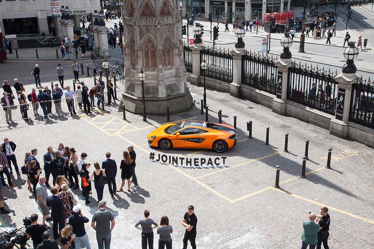 Crowd gathered around s sports car with a banner showing "Join the pact" outside of a train station