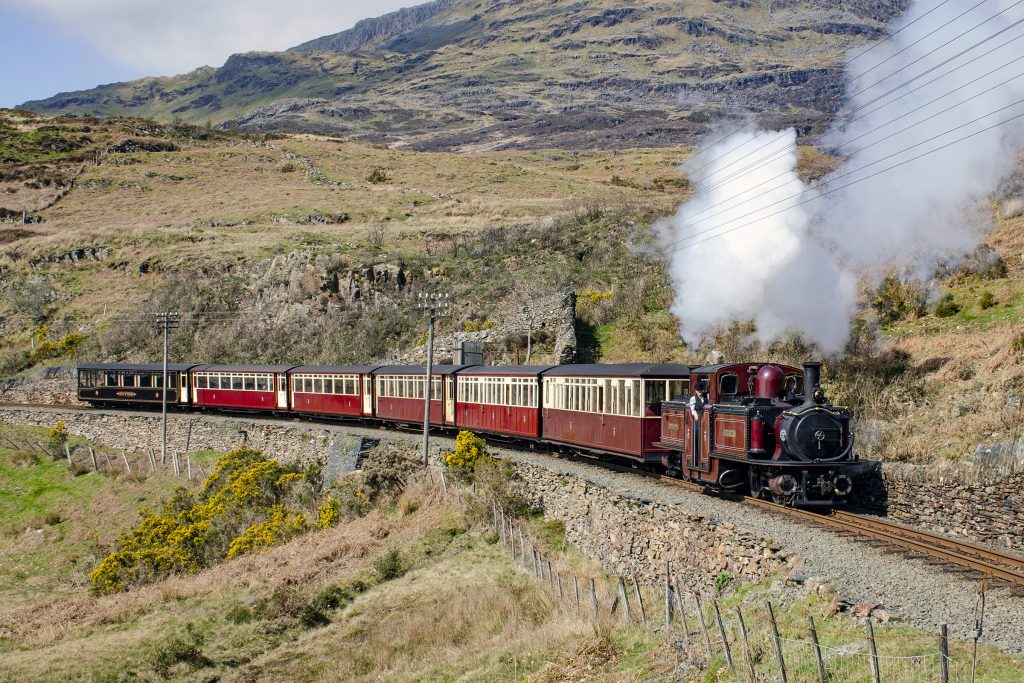 A steam train travels along the Ffestiniog Railway during the daytime in North Wales with mountains in the background