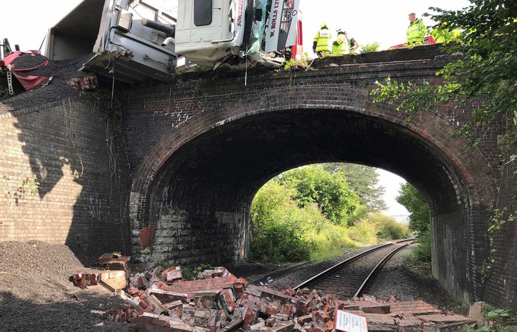 A lorry on it's side that has crashed over a bridge, with emergency workers at the scene