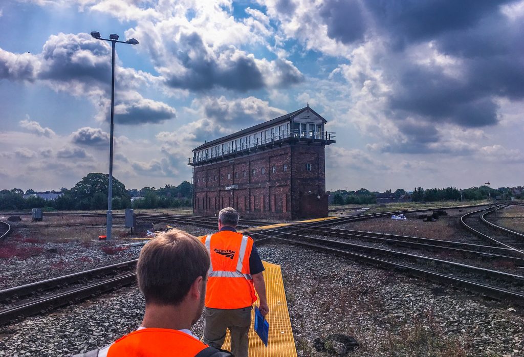 Landscape view of the three-storey Severn Bridge Junction signal box in the middle of the tracks with two people in PPE in the foreground