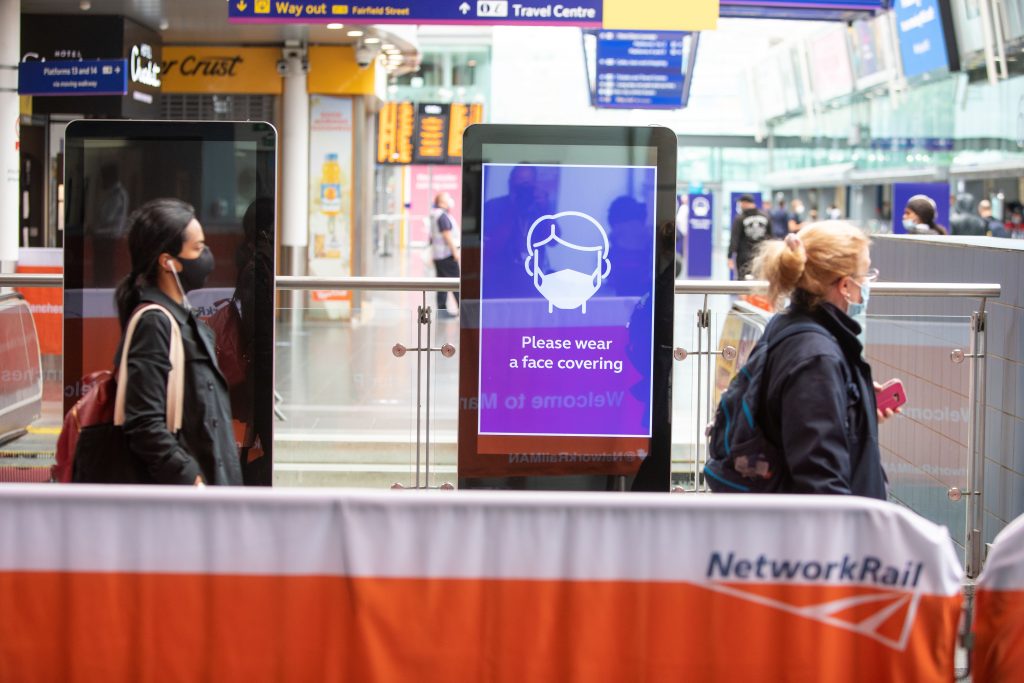 Passengers wearing face coverings walk past a face coverings image on an advertising screen in a station