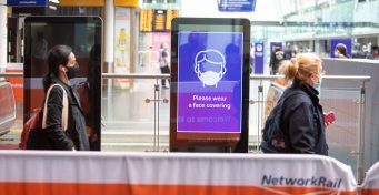Passengers wearing face coverings walk past a face coverings image on an advertising screen in a station