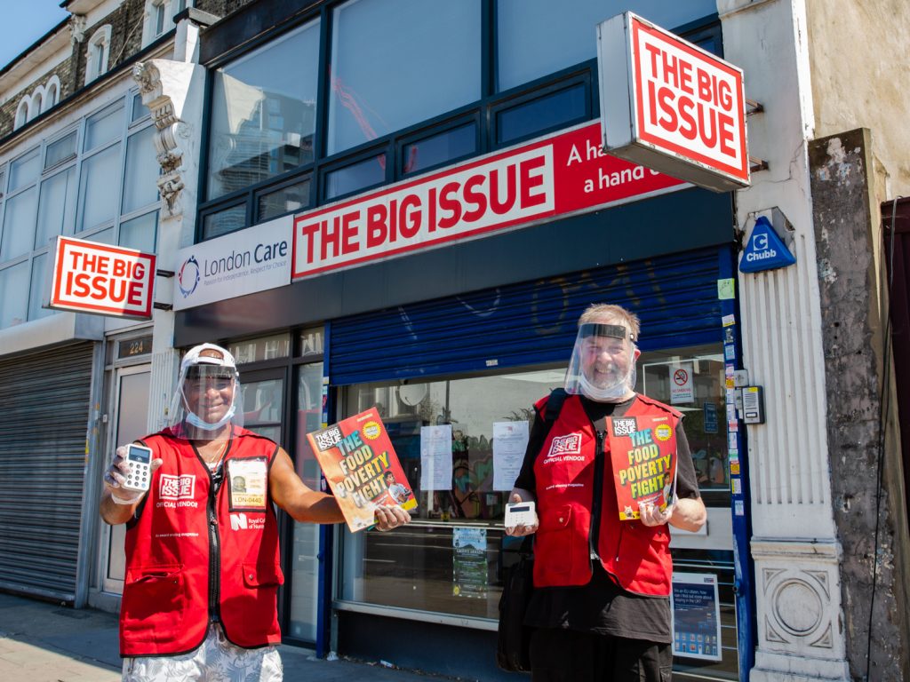 Two Big Issue vendors wearing covid protection, holding The Big Issue magazines and a card payment machine.
