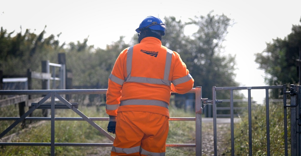 Nathaniel, a Network Rail ecologist standing against a gate in full PPE, looking into a field