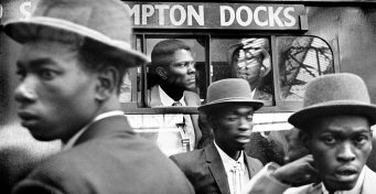 1960s photograph of the Windrush Generation arriving at London Waterloo - men in smart hats in front of a bus.