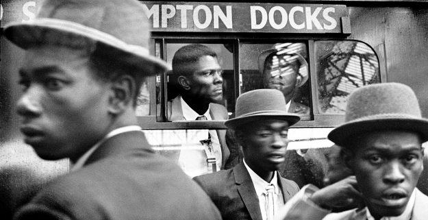 1960s photograph of the Windrush Generation arriving at London Waterloo - men in smart hats in front of a bus.