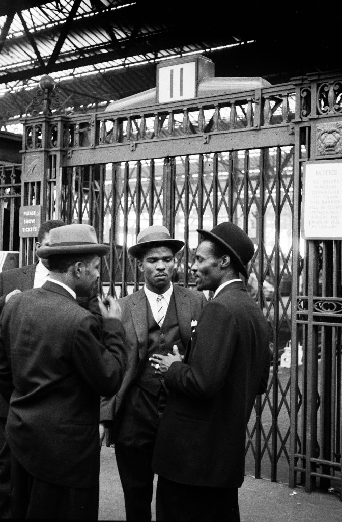 1960s photograph of the Windrush Generation arriving at London Waterloo - men in smart hats and suits talking at a railway station