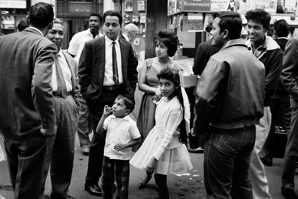 1960s photograph of the Windrush Generation arriving at London Waterloo - men, women and children, standing in a circle talking and smiling