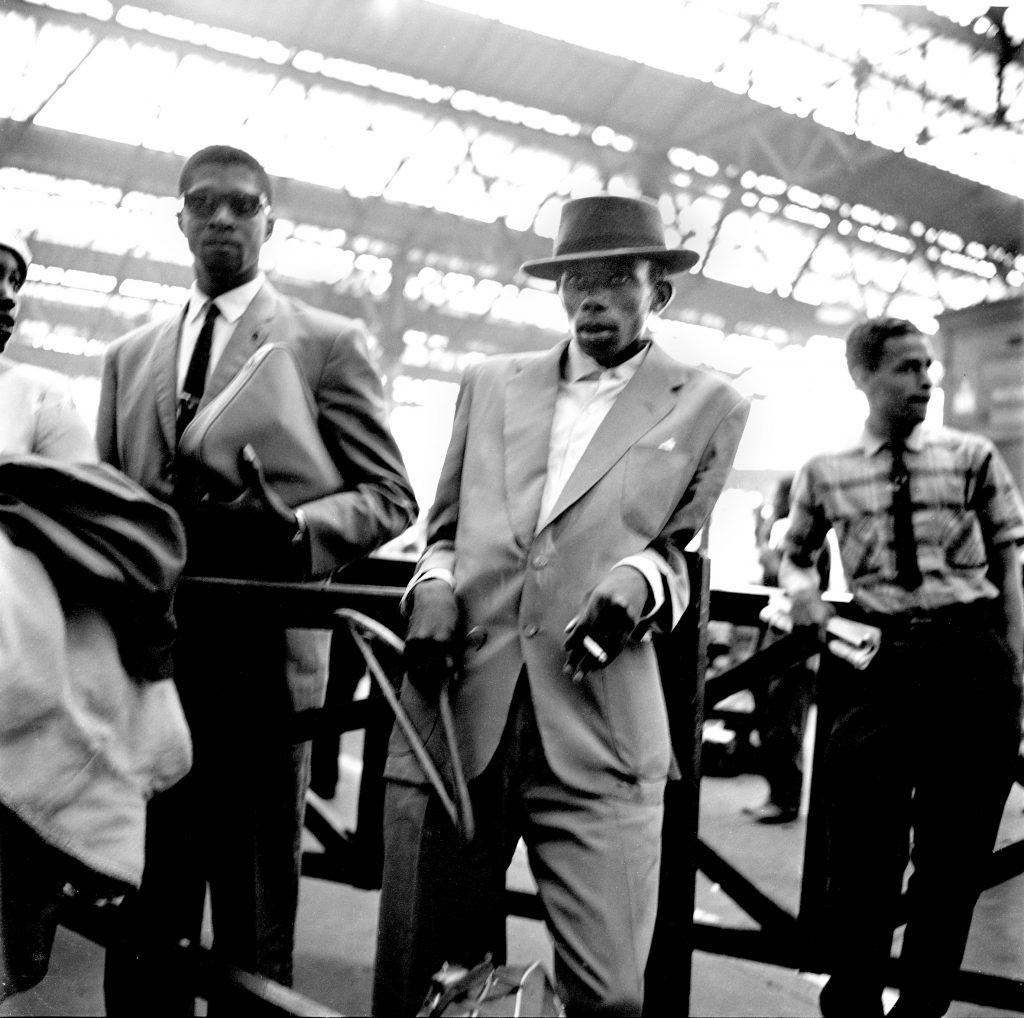 1960s photograph of the Windrush Generation arriving at London Waterloo - a smartly dressed man leans against some railings
