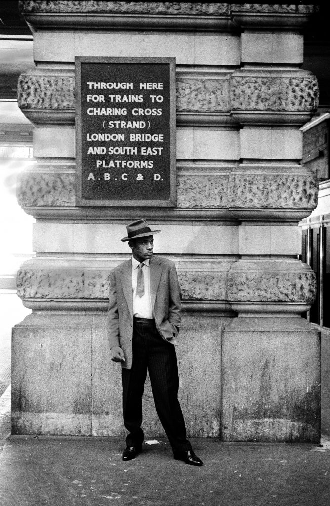 A black man standing at a railway station under a sign providing platform information
