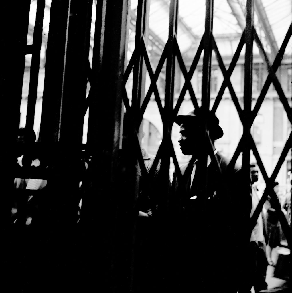 A 1960s photograph of a smartly dressed member of the windrush generation behind railway railings