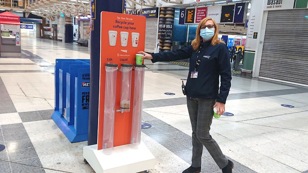 A station staff member demonstrating the new coffee cup recycling bins on a station concourse, daytime.