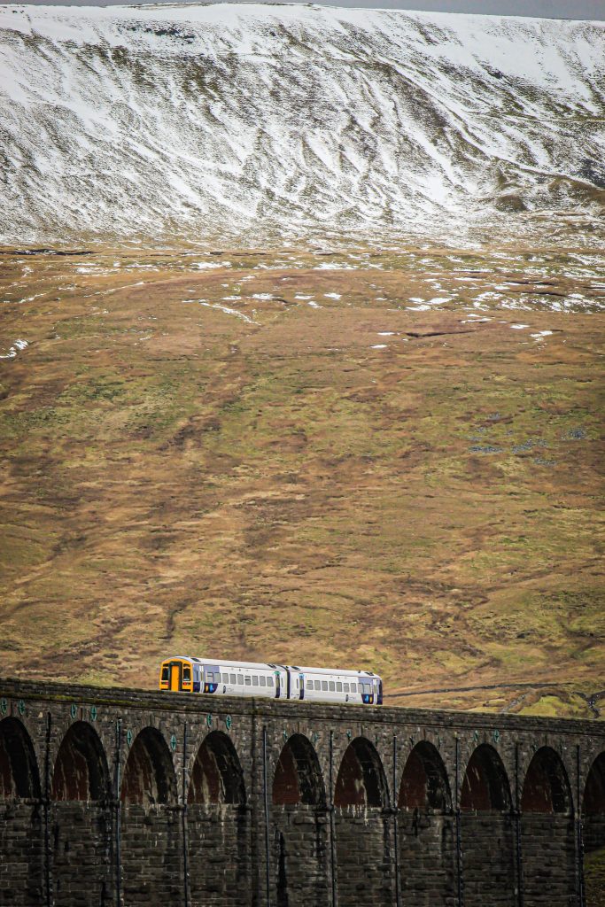 A train crosses the Ribblehead Viaduct with snowy peaks in the background, daytime