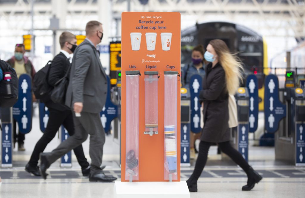 Passengers walk past a coffee cup recycling bin on a station concourse.