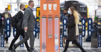 Passengers walk past a coffee cup recycling bin on a station concourse.