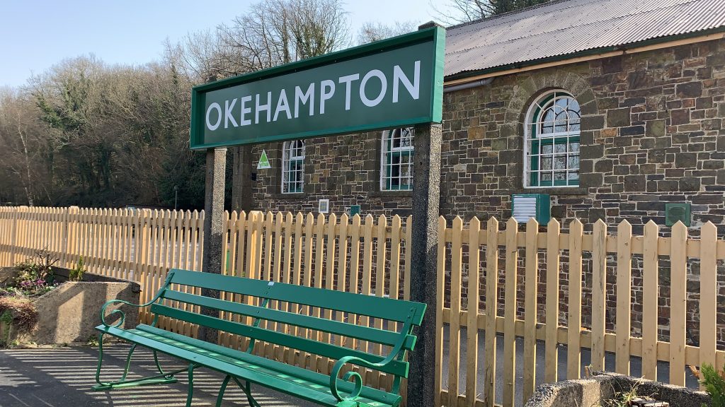 A green bench beneath the Okehampton sign on the platform at Okehampton railway station, daytime