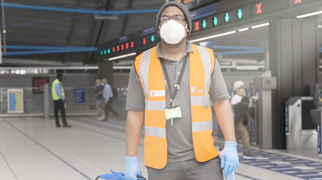 A station staff member in orange vest, face mask and plastic gloves with ticket barriers in background, daytime