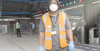 A station staff member in orange vest, face mask and plastic gloves with ticket barriers in background, daytime
