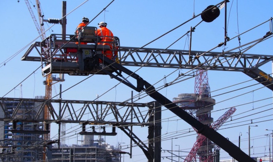 Two railway workers attend to overhead line equipment with the ArcelorMittal Orbit sculpture in background, daytime