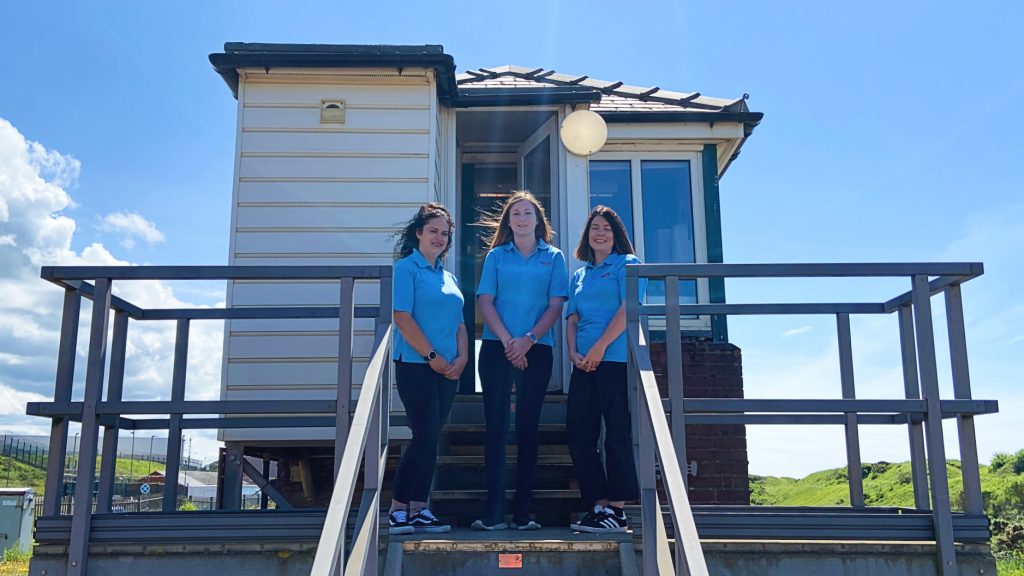 A team of three female signallers stands outside a small, traditional signal box, daytime