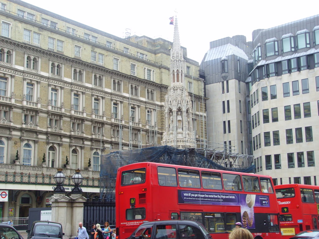 A red London bus drives past the restored Eleanor Cross, daytime