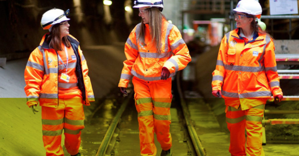 Three female Level 3 engineering apprentices in orange Network Rail workwear chat happily walking through a railway tunnel.