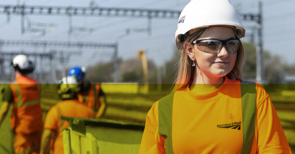 Female rail graduate in orange Network Rail uniform and hard hat gazes across rail tracks.