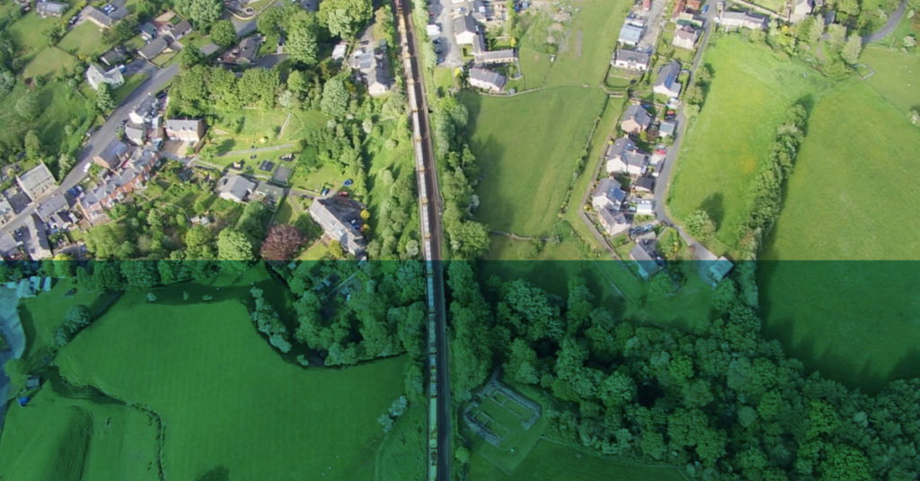 Bird's eye view of a train track running through a countryside environment with trees, fields and houses either side.