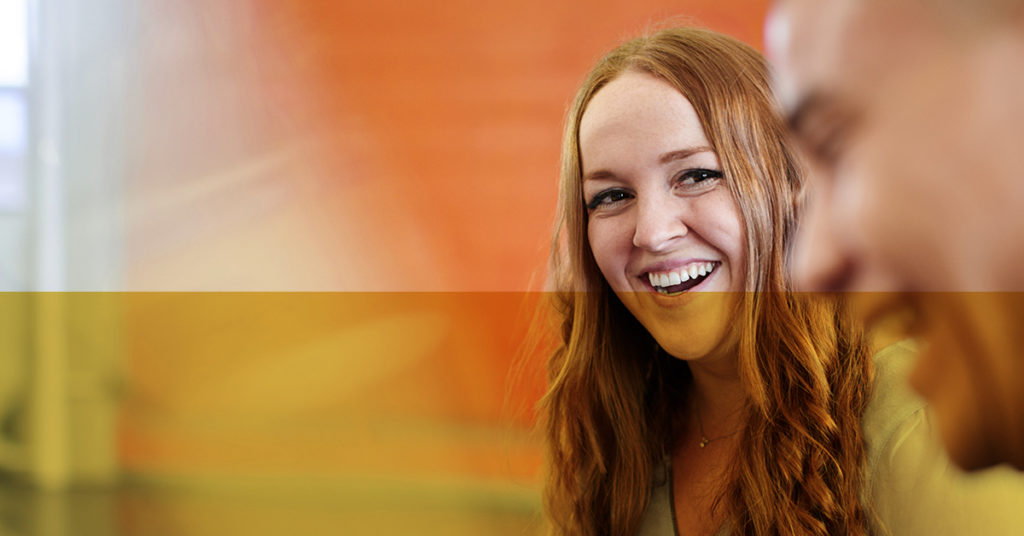 Female rail graduate with long wavy hair smiles at her male colleague in the foreground against an orange wall.