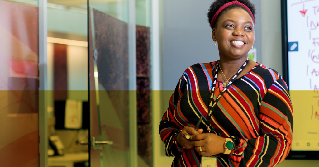 Female operations management graduate in stripy top smiles proudly in glass office environment.