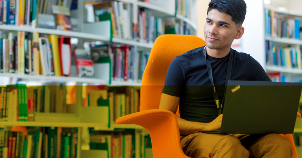 Male rail graduate sits on orange chair in a library gazing away from his laptop.