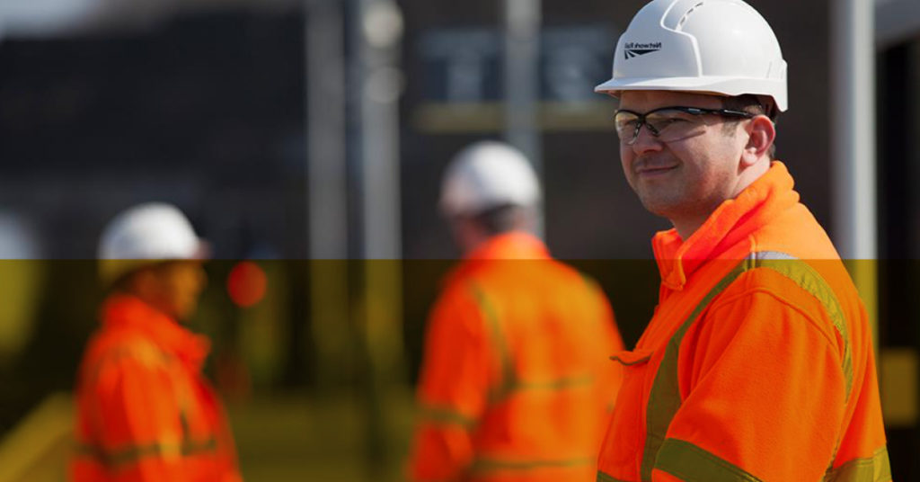 Male mechanical engineering graduate in hard hat and orange jacket gazes off camera.