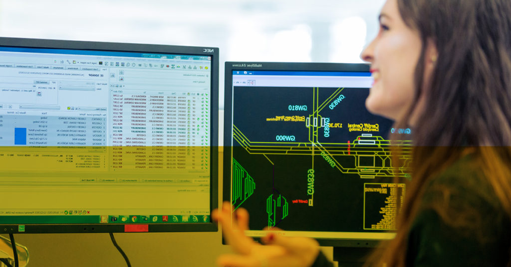 Female strategy planning graduate smiles at computer screens showing a rail system and spreadsheet.