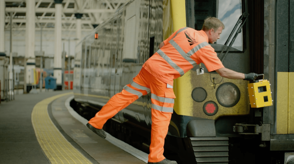 A Network Rail employee in PPE fitting an AIVR-Go camera to the front of a train at a railway station.