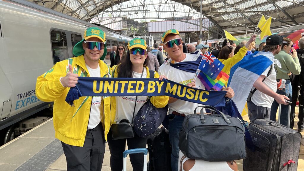 Eurovision fans on a platform at Liverpool Lime Street holding a banner that reads 'united by music'.