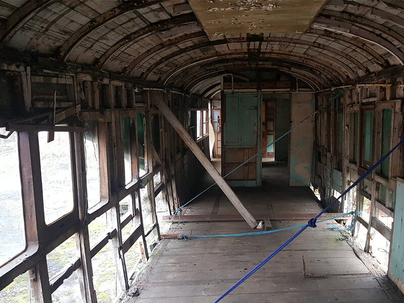 Interior of an old second/third class compartment of a Titanic boat train showing worn wooden floors and exposed metal framework, with doors and windows visible along the sides.